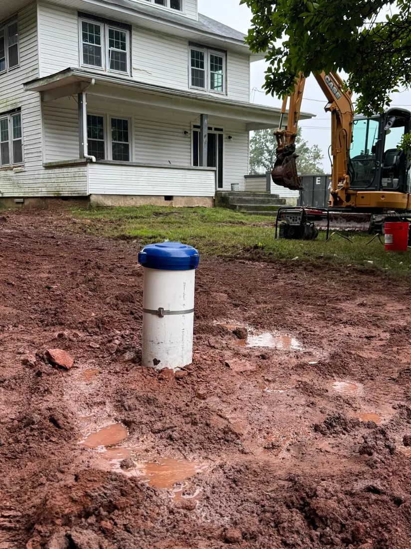 Capped well casing in a red clay yard beside an older Maryland farmhouse, with an excavator at work